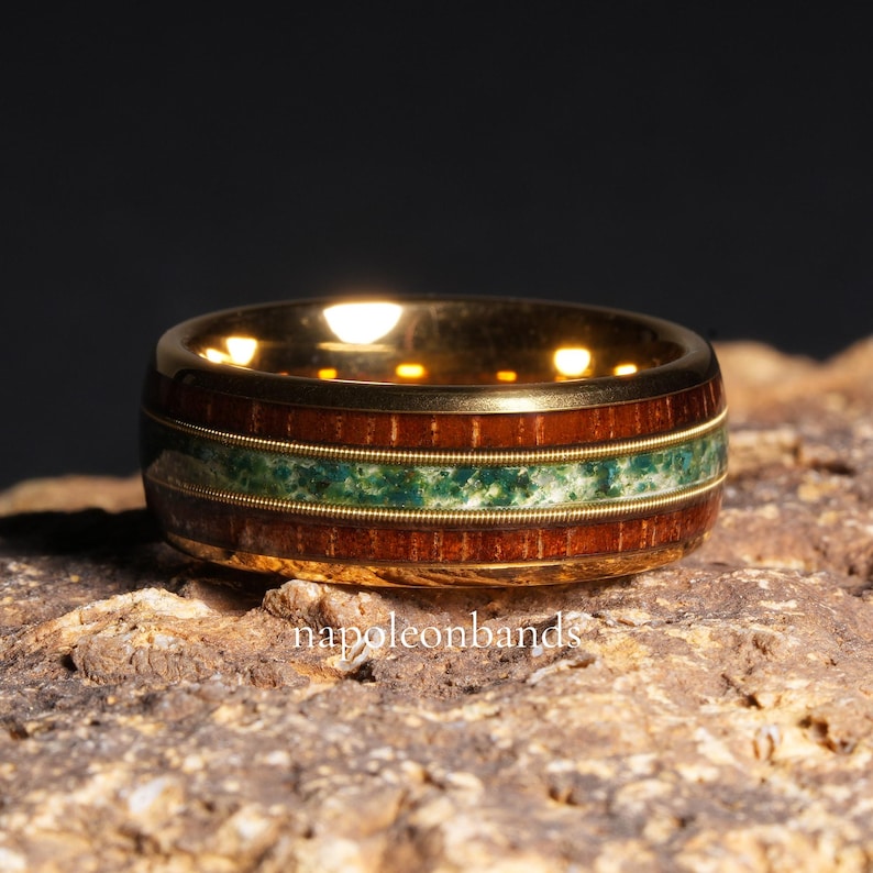 May include: A gold-colored ring with a band of dark wood, a central stripe of green stone, and thin gold bands. The ring is set on a textured, brown surface. The text "napoleonbands" is visible.