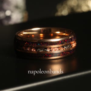 May include: A rose gold ring with a dark inlay featuring red and black flecks, and a band of copper-colored material. The ring has a polished finish and the text "napoleonbands" is visible.