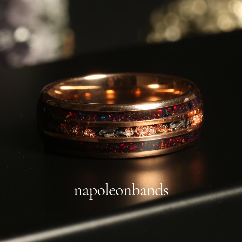 May include: A rose gold ring with a dark inlay featuring red and black flecks, and a band of copper-colored material. The ring has a polished finish and the text "napoleonbands" is visible.