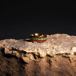 May include: A gold-colored ring with a wooden band and a green stone inlay. The ring features a dark wood grain and a band of small green stones. The text "napoleonbands" is visible on the surface below the ring.