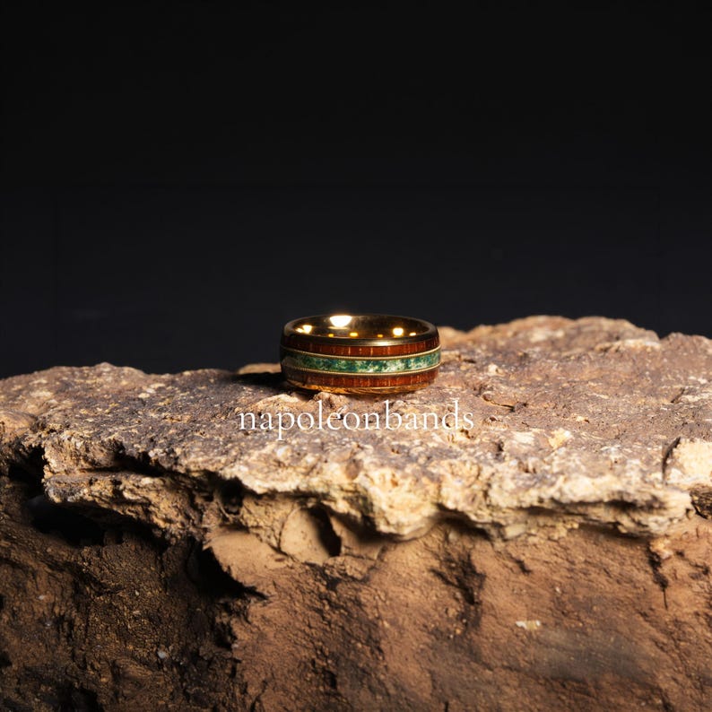 May include: A gold-colored ring with a wooden band and a green stone inlay. The ring features a dark wood grain and a band of small green stones. The text "napoleonbands" is visible on the surface below the ring.