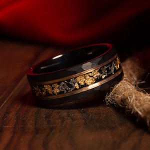 May include: Black ring with a hammered texture, featuring a gold-colored inlay with a textured design. The ring has a dark, metallic appearance, with a band of gold and silver-colored accents. The ring is set against a dark background.