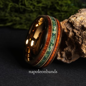 May include: A gold-colored ring with a wood and stone inlay. The ring features a band of reddish-brown wood, a strip of green stone, and a thin gold band. The text "napoleonbands" is at the bottom of the image.