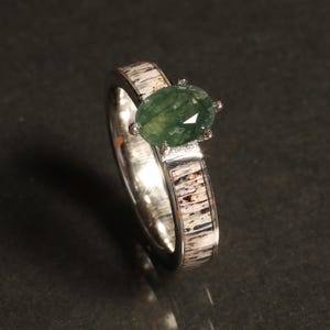 May include: A silver ring with a green gemstone set in a prong setting. The band is made of antler and has a natural wood grain pattern.
