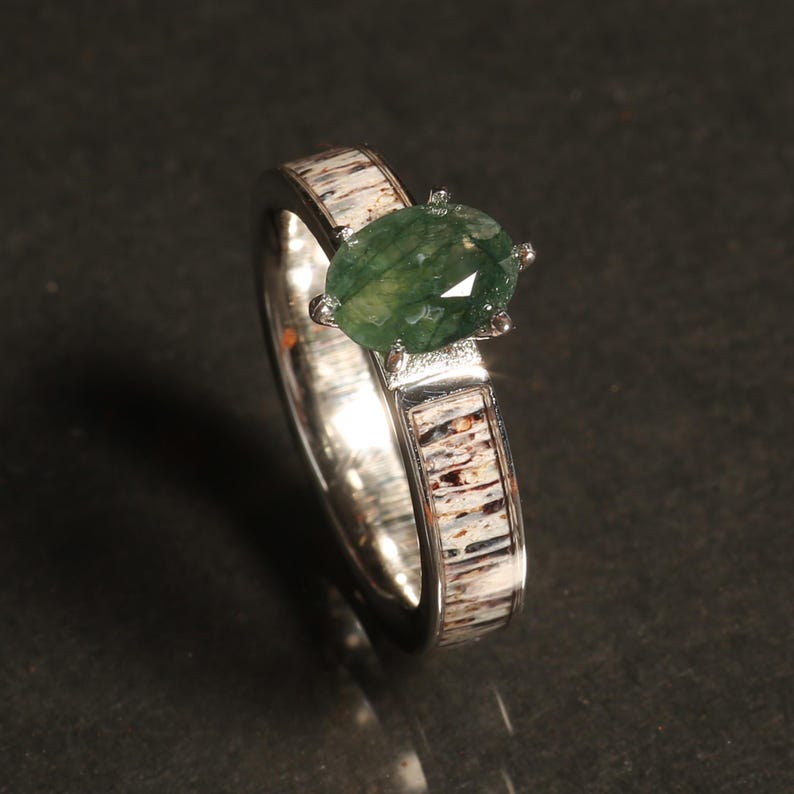 May include: A silver ring with a green gemstone set in a prong setting. The band is made of antler and has a natural wood grain pattern.