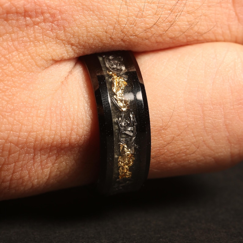 May include: Black ring with gold flakes and a clear resin inlay. The ring is on a finger. The band is black and has a polished finish. The gold flakes are irregularly shaped and add a touch of sparkle.