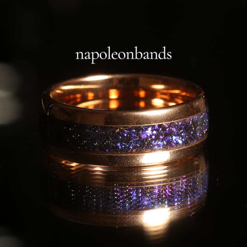 May include: A rose gold ring with a band of sparkling purple and blue material. The ring is reflected in the dark surface below. The text "napoleonbands" is displayed in white above the ring.