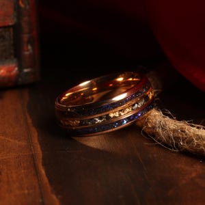 Puede incluir: Un anillo de oro rosa con incrustaciones azules y doradas. El anillo tiene un acabado pulido y se encuentra sobre un fondo de madera.
