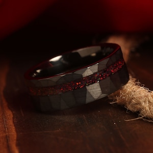 May include: A black tungsten carbide ring with a red and black inlay. The ring has a hammered finish and a thin red line around the band.