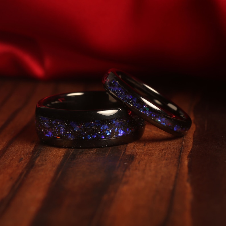 May include: Two black wedding bands with a blue glitter inlay. The bands are on a wooden surface.