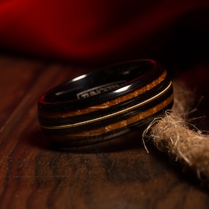 May include: A black tungsten ring with a brown wood inlay and two gold bands. The ring is on a wooden surface.