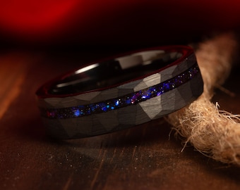 May include: A black ring with a textured surface and a band of sparkling blue and purple stones. The ring is set against a dark background, highlighting its intricate design and colour. The ring is a piece of jewellery.