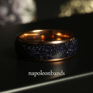 May include: A close-up of a ring with a dark blue, glittery band and a gold-colored interior and rim. The ring is set against a dark background, with the text "napoleonbands" at the bottom.