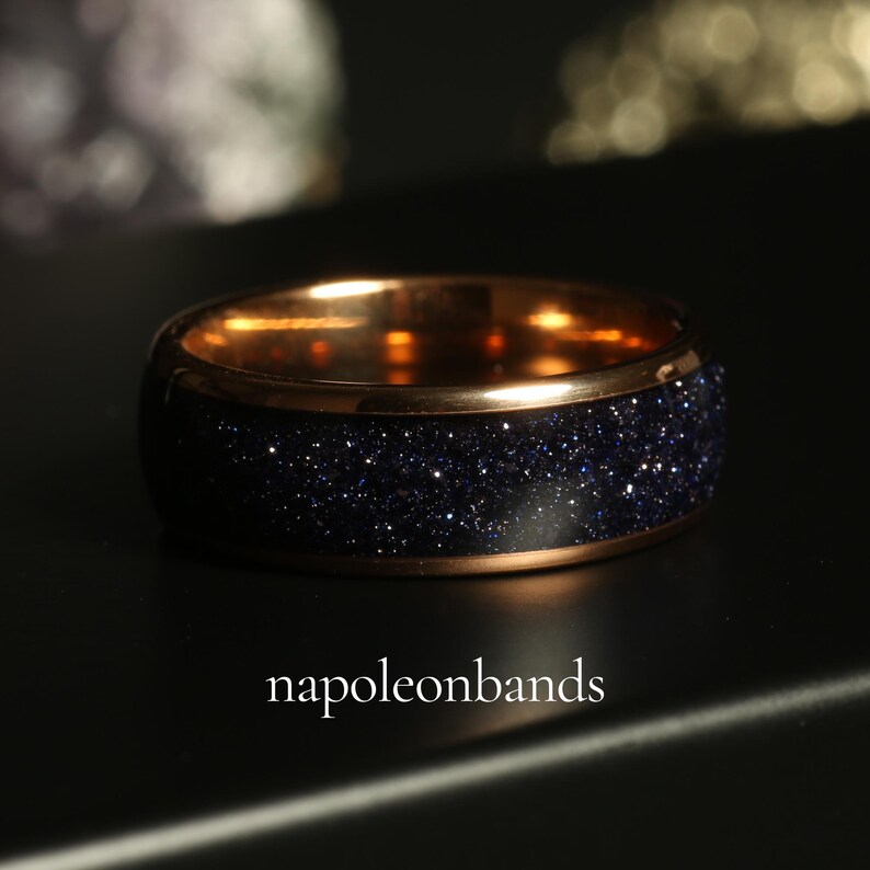 May include: A close-up of a ring with a dark blue, glittery band and a gold-colored interior and rim. The ring is set against a dark background, with the text "napoleonbands" at the bottom.