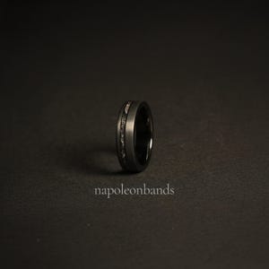 May include: A black tungsten carbide ring with a thin silver inlay. The ring is on a black background.