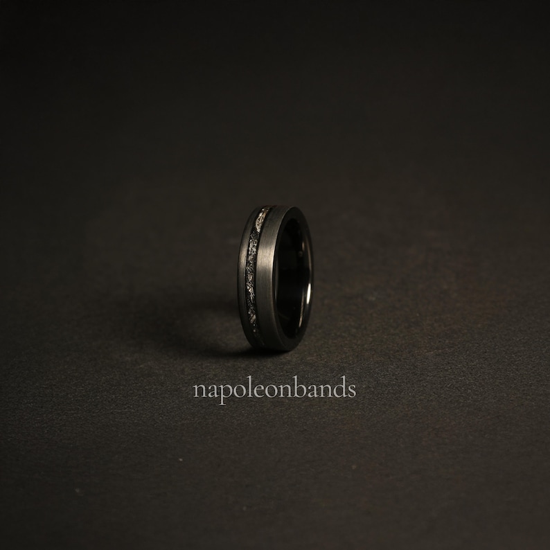 May include: A black tungsten carbide ring with a thin silver inlay. The ring is on a black background.