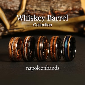 May include: Five different men's wedding bands with a whiskey barrel theme. The bands are made of black tungsten carbide with various wood inlays. The text "Whiskey Barrel Collection" is displayed above the rings. The text "napoleonbands" is displayed below the rings.