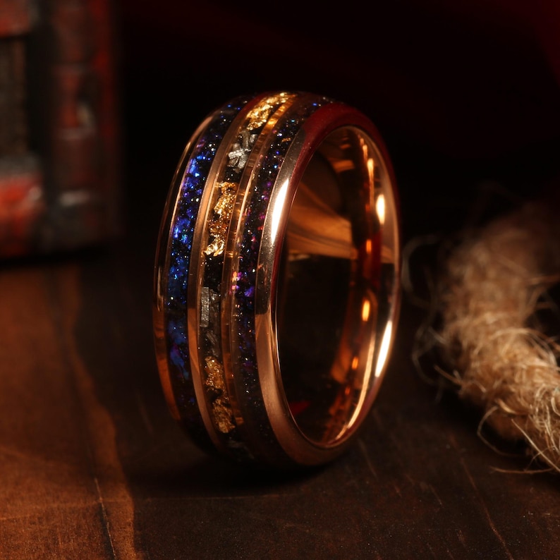 Puede incluir: Un anillo de carburo de tungsteno de color oro rosa con una incrustaci&oacute;n azul y dorada. La incrustaci&oacute;n presenta un patr&oacute;n de motas doradas y brillo azul.