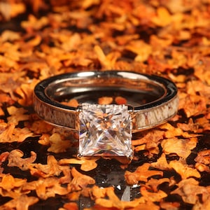 Puede incluir: Un anillo de plata con una piedra preciosa transparente de forma cuadrada engastada en el centro. La banda del anillo está hecha de asta de ciervo.
