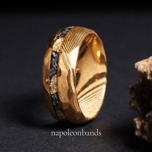 May include: A gold-colored ring with a textured surface and a band of black stones. The ring has a unique design with diagonal lines and a hammered effect. The text "napoleonbands" is at the bottom of the image.