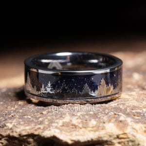 May include: A silver-toned ring with a dark blue, starry inlay featuring a silhouetted forest scene. The ring has a polished finish and a band that is approximately 8mm wide. The text "napoleonbands" is visible on the ring.