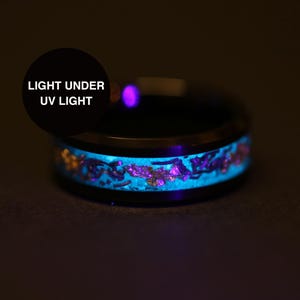May include: Black ring with a glowing blue and purple inlay. The ring's design includes gold accents. The text "LIGHT UNDER UV LIGHT" is displayed in a black circle. The ring is shown in a dark setting.