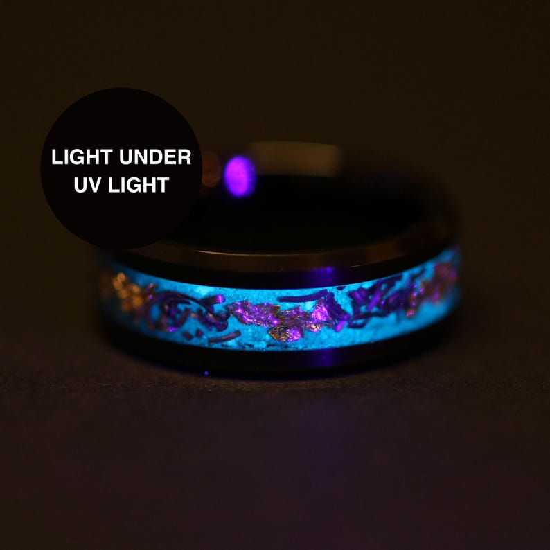 May include: Black ring with a glowing blue and purple inlay. The ring's design includes gold accents. The text "LIGHT UNDER UV LIGHT" is displayed in a black circle. The ring is shown in a dark setting.