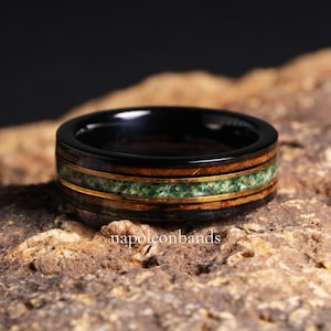 Puede incluir: Un anillo negro con una banda de vetas de madera, una incrustación de piedra verde y detalles dorados. El anillo está sobre una superficie marrón texturizada. El texto "napoleonbands" es visible.