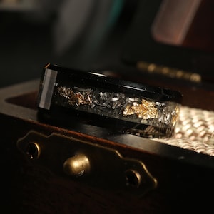 May include: A black ring with a band of gold and silver flakes encased in clear resin. The ring is set against a dark background, highlighting the contrast of the materials and colors. The ring is displayed in a wooden box.