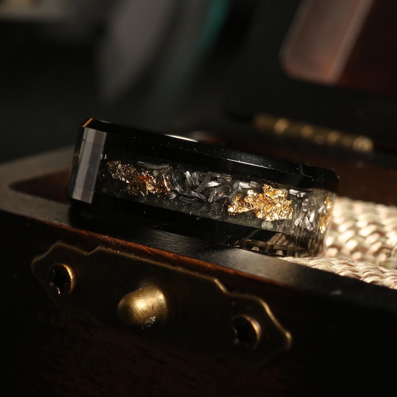 May include: A black ring with a band of gold and silver flakes encased in clear resin. The ring is set against a dark background, highlighting the contrast of the materials and colors. The ring is displayed in a wooden box.