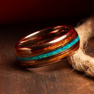 May include: A rose gold ring with a wooden inlay and a blue opal accent. The ring is sitting on a wooden surface.