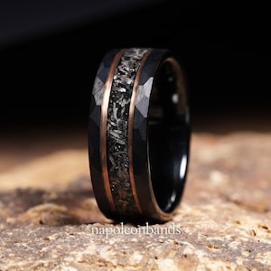 May include: Close-up of a black ring with a hammered texture. The ring features a central band of dark gray material, framed by thin copper-colored bands. The text "napoleonbands" is visible at the bottom of the image. The ring is set on a light brown surface.