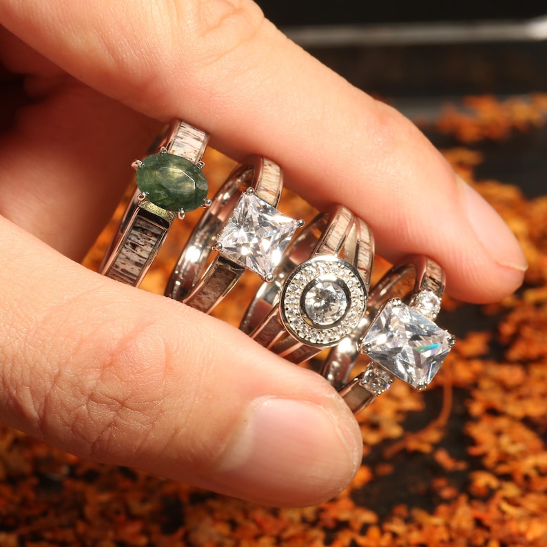 May include: A stack of five rings with different stones. The top ring has a green stone, the next ring has a clear square stone, the next ring has a clear round stone with a silver halo, and the bottom two rings have clear rectangular stones.