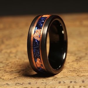 Puede incluir: Un anillo de carburo de tungsteno negro con una incrustación de cobre y azul. La incrustación presenta un patrón de escamas de cobre y resina azul.