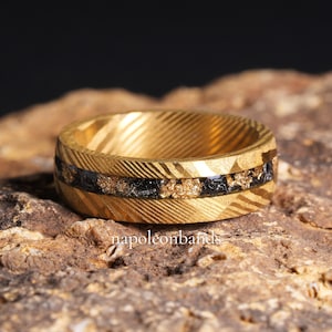 May include: A gold ring with a textured pattern and a black and gold inlay. The ring features a band of black material with gold flecks. The ring is displayed on a brown surface. The text "napoleonbands" is visible.