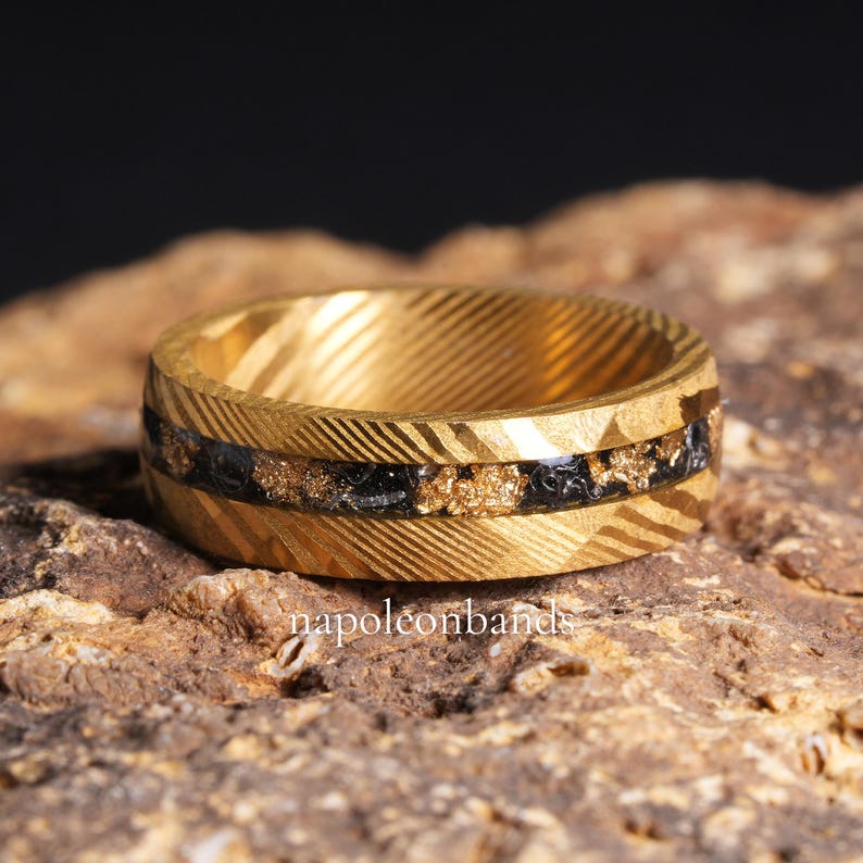 May include: A gold ring with a textured pattern and a black and gold inlay. The ring features a band of black material with gold flecks. The ring is displayed on a brown surface. The text "napoleonbands" is visible.