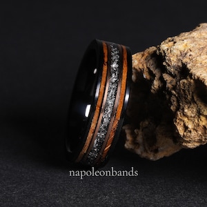 May include: A black ring with a wood grain and silver inlay. The ring is next to a rough, tan rock. The text "napoleonbands" is at the bottom of the image.