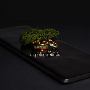 May include: Two gold-colored rings with wood and green stone inlays. One ring is stacked on top of the other. The rings are on a black reflective surface with green moss in the background. The text "napoleonbands" is at the bottom.