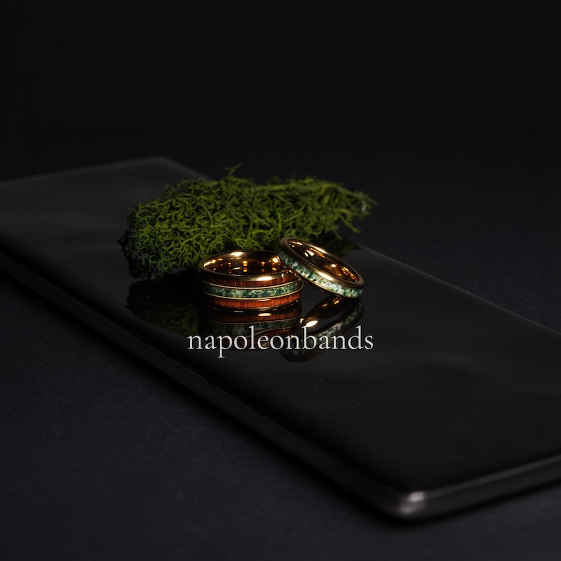 May include: Two gold-colored rings with wood and green stone inlays. One ring is stacked on top of the other. The rings are on a black reflective surface with green moss in the background. The text "napoleonbands" is at the bottom.
