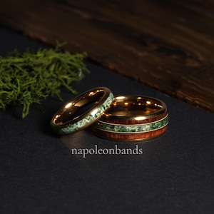 May include: Two gold-colored rings. One ring features a band of green stone, while the other has a band of green stone between two bands of wood. The rings are on a black surface. The text "napoleonbands" is visible.
