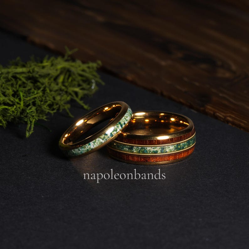 May include: Two gold-colored rings. One ring features a band of green stone, while the other has a band of green stone between two bands of wood. The rings are on a black surface. The text "napoleonbands" is visible.