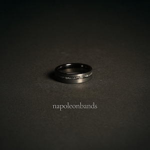 May include: A black tungsten carbide ring with a thin band of silver inlay. The ring is on a dark gray surface.