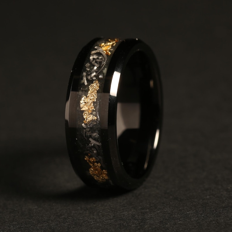May include: A black ring with a gold-colored inlay. The ring has a glossy finish and a band of gold flakes and intricate detailing. The ring is set against a dark background, highlighting its reflective surface.