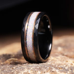 May include: A black ring with a hammered texture and a central band of white opal. The opal is flanked by thin bands of rose gold. The ring is set against a dark background, highlighting its details.