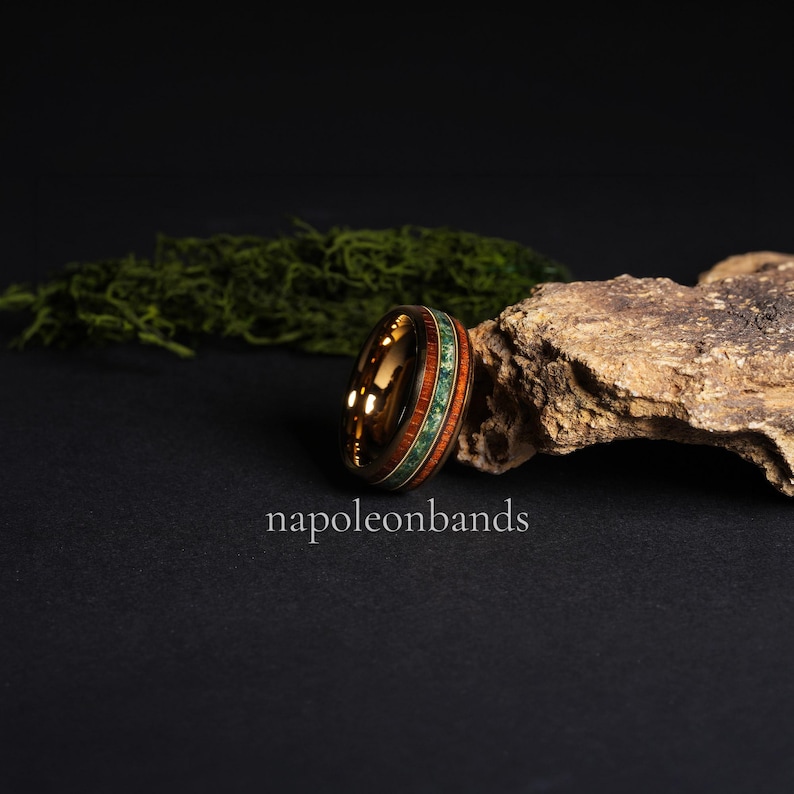 May include: A gold-colored ring with a wood and green stone inlay. The ring features a polished finish and is set against a dark background with the text "napoleonbands". The ring's design incorporates natural elements.