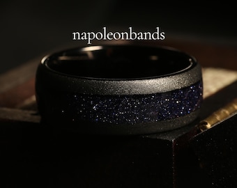 May include: A black ring with a band of blue glitter embedded in the metal. The ring is on a dark surface. The text "napoleonbands" is visible above the ring.