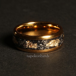 May include: A gold and black ring with a band of black metal and gold flecks. The ring is on a black surface.