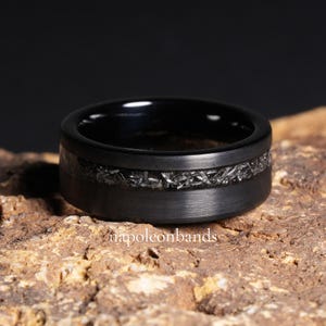 Peut inclure: Une bague noire avec une finition en métal brossé et une bande de matériau texturé argenté. La bague est posée sur une surface rugueuse et brune. Le texte "napoleonbands" est visible.