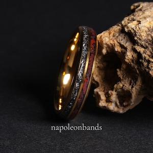 May include: A gold-colored ring with a black and red inlay. The ring is set against a dark background and a piece of textured rock. The text "napoleonbands" is visible at the bottom of the image.
