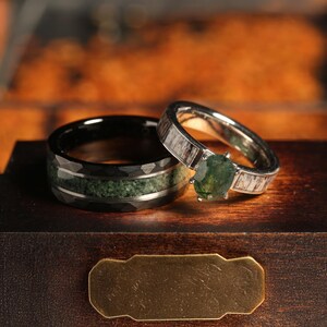 May include: A pair of wedding bands, one with a black and green inlay and the other with a green gemstone set in a silver band with a wood-like pattern.